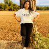 Mujer con la camiseta cristiana GRÀCIA en color crudo, apoyada en un árbol en un campo al atardecer