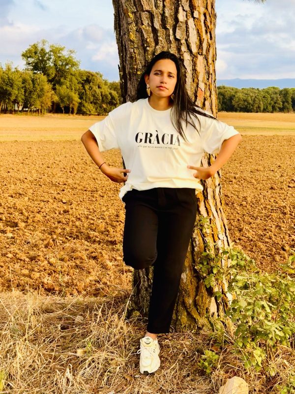 Mujer con la camiseta cristiana GRÀCIA en color crudo, apoyada en un árbol en un campo al atardecer
