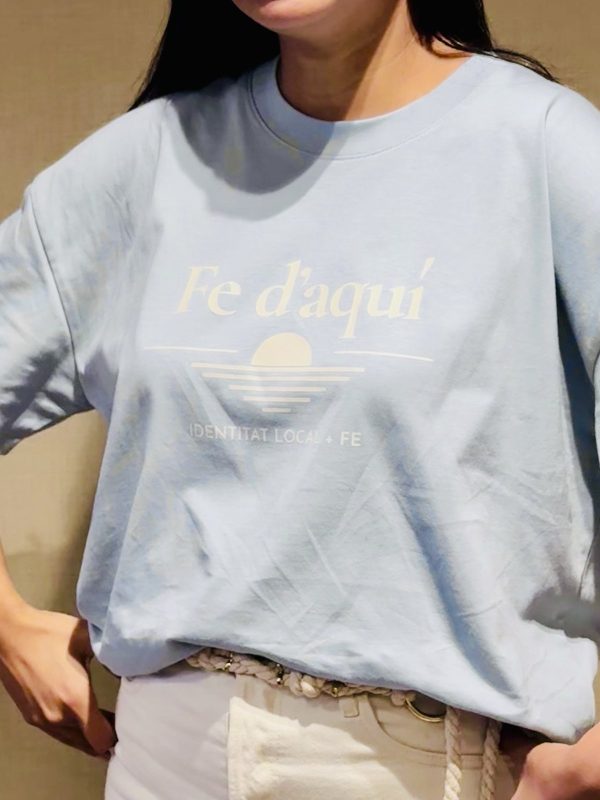 Camiseta Fe d'aquí azul — detalle del estampado Detalle del estampado Fe d'aquí, identitat local + fe, sobre camiseta azul mediterráneo
