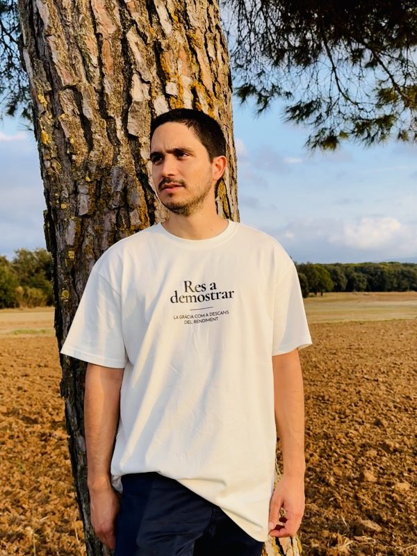 Camiseta Res a demostrar — hombre en el campo Hombre con la camiseta cristiana Res a demostrar en color crudo, apoyado en un pino
